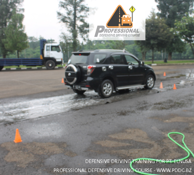 Seorang peserta pelatihan mengendarai motor dan mempraktikkan teknik pengereman darurat di permukaan jalan berpasir atau berkerikil, dengan instruktur mengawasi di area pelatihan PDDC.